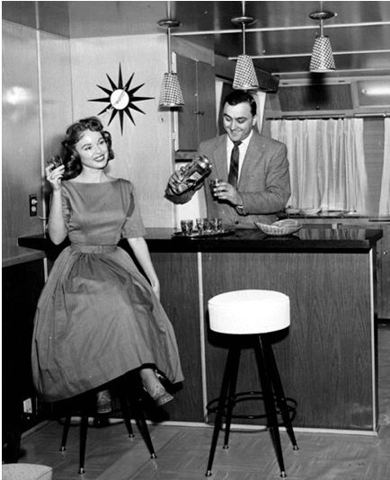 1950s-vintage-photo-of-a-woman-in-a-1950s-dress-sitting-on-a-bar-stool-at-her-home-bar-while-her-husband-makes-a-drink-behind-the-bar.jpg